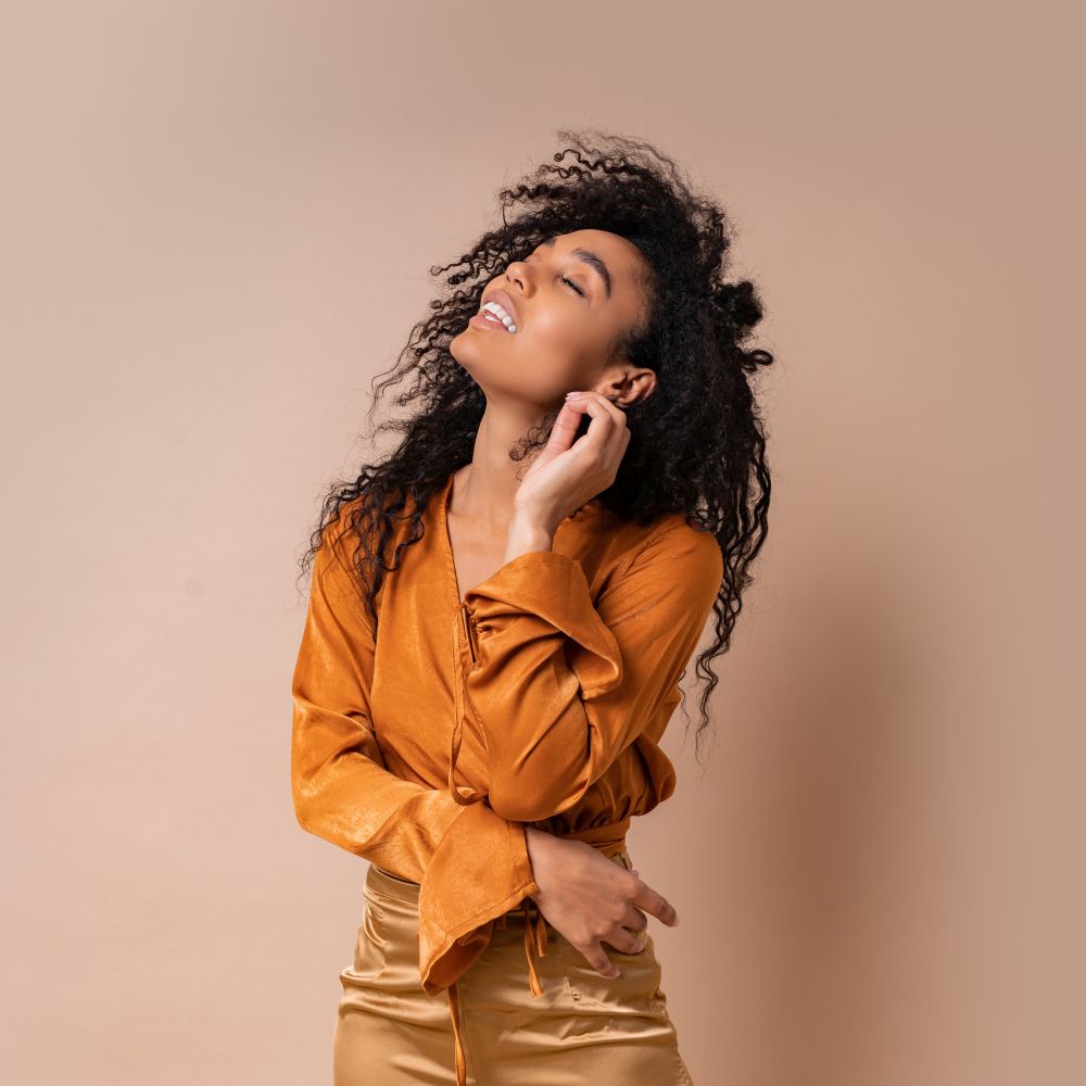 Glad african woman with perfect curly hairs in casual orange blouse and golden pants posing on beige background.