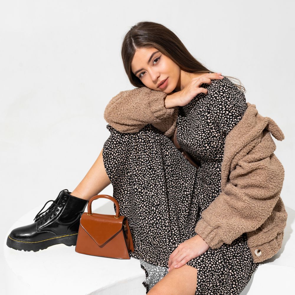 Graceful european woman in winter fur coat and stylish dress  sitting in studio over white background. Wearing  ankle boot in black leather.