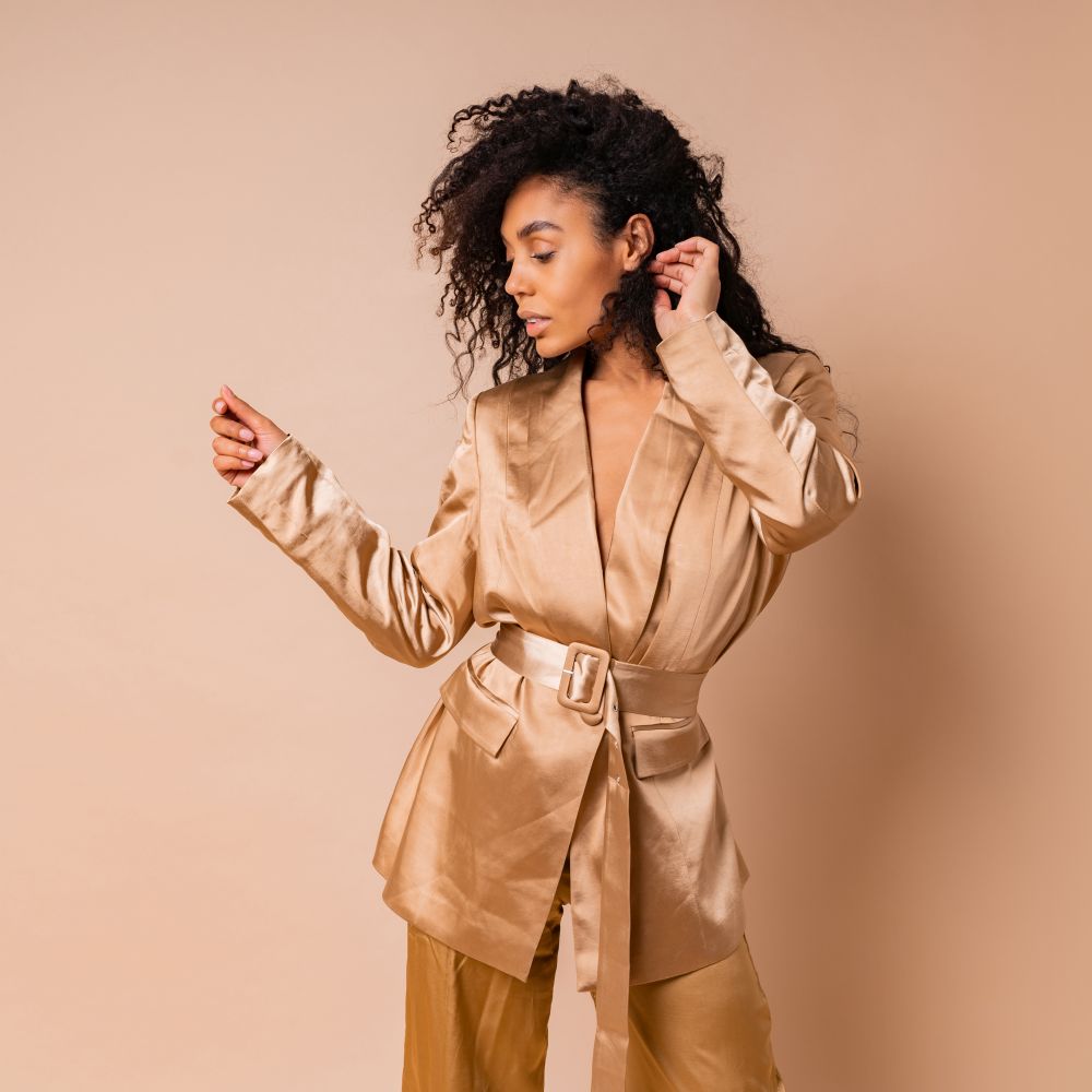 Sensual black woman with beautiful wavy hairs in elegant  golden satin suit posing over beige background. Spring fashion look.