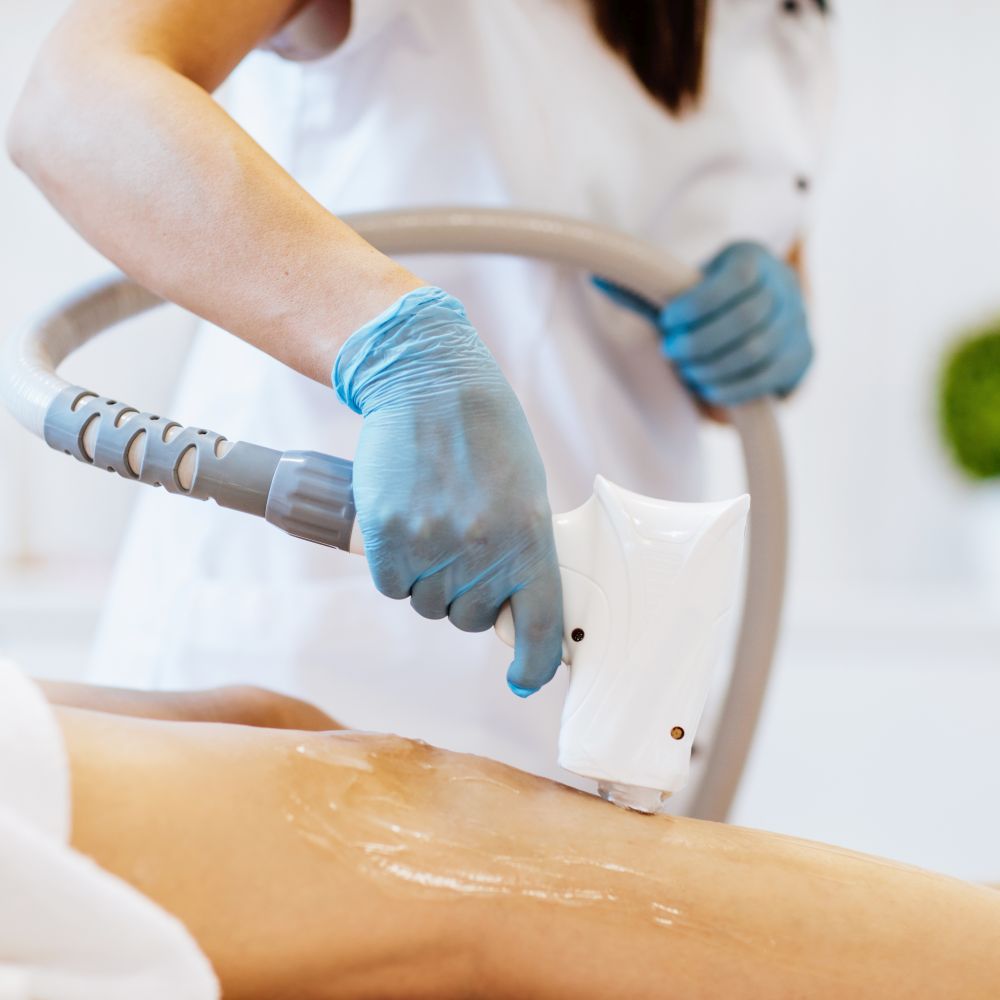 Doctors's hands in blue medical gloves holding a depilation machine and using it on the woman's legs. Focus is at the doctor's hands.Copy space.